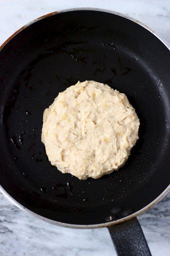 Raw banana pancake in a black frying pan against a marble background