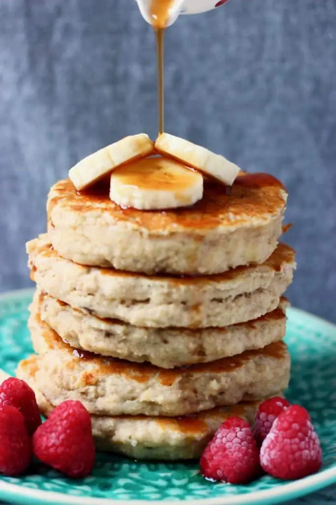 Stack of five banana pancakes topped with banana slices with maple syrup being poured over the top on a green plate with raspberries against a grey background