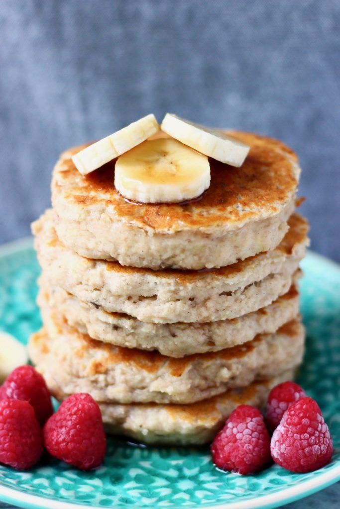 Stack of five banana pancakes topped with banana slices on top on a green plate with raspberries against a grey background