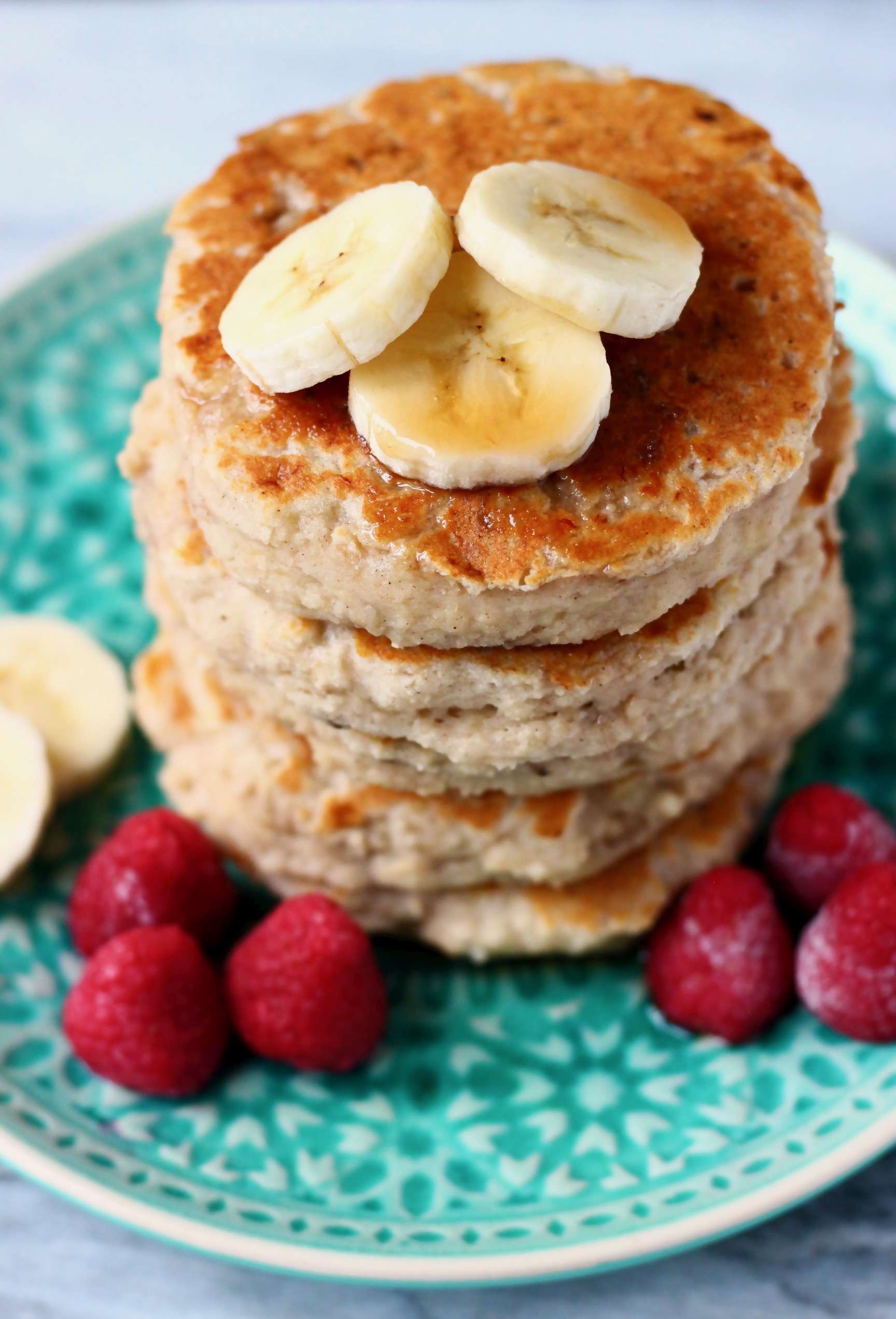 Stack of five banana pancakes topped with banana slices on top on a green plate with raspberries against a marble background