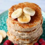 Stack of five banana pancakes topped with banana slices on top on a green plate with raspberries against a marble background