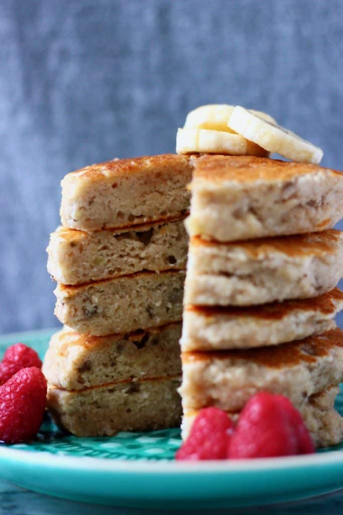 Stack of five banana pancakes topped with banana slices on top on a green plate with raspberries against a grey background