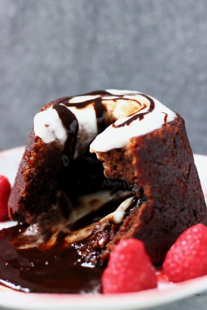 Chocolate lava cake on a plate with chocolate sauce flowing out of it topped with white cream and decorated with raspberries against a grey background