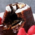 Gluten-free vegan chocolate lava cake on a plate with chocolate sauce flowing out of it topped with white cream and decorated with raspberries