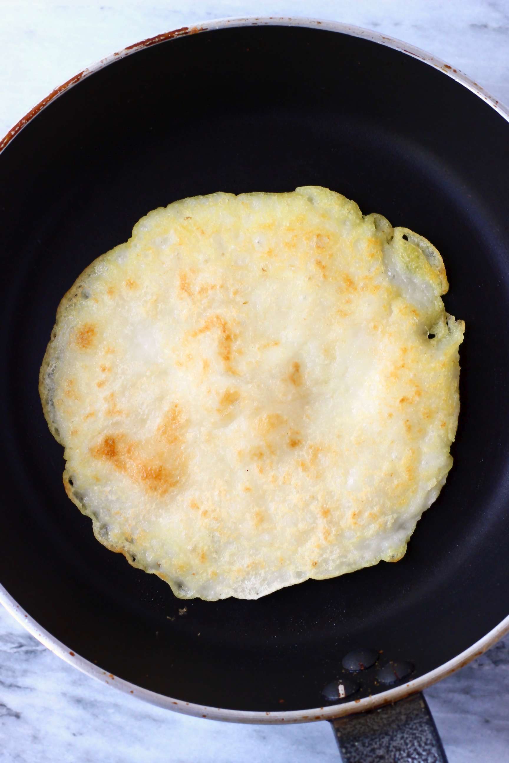 A round gluten-free vegan crepe being fried in a black frying pan 
