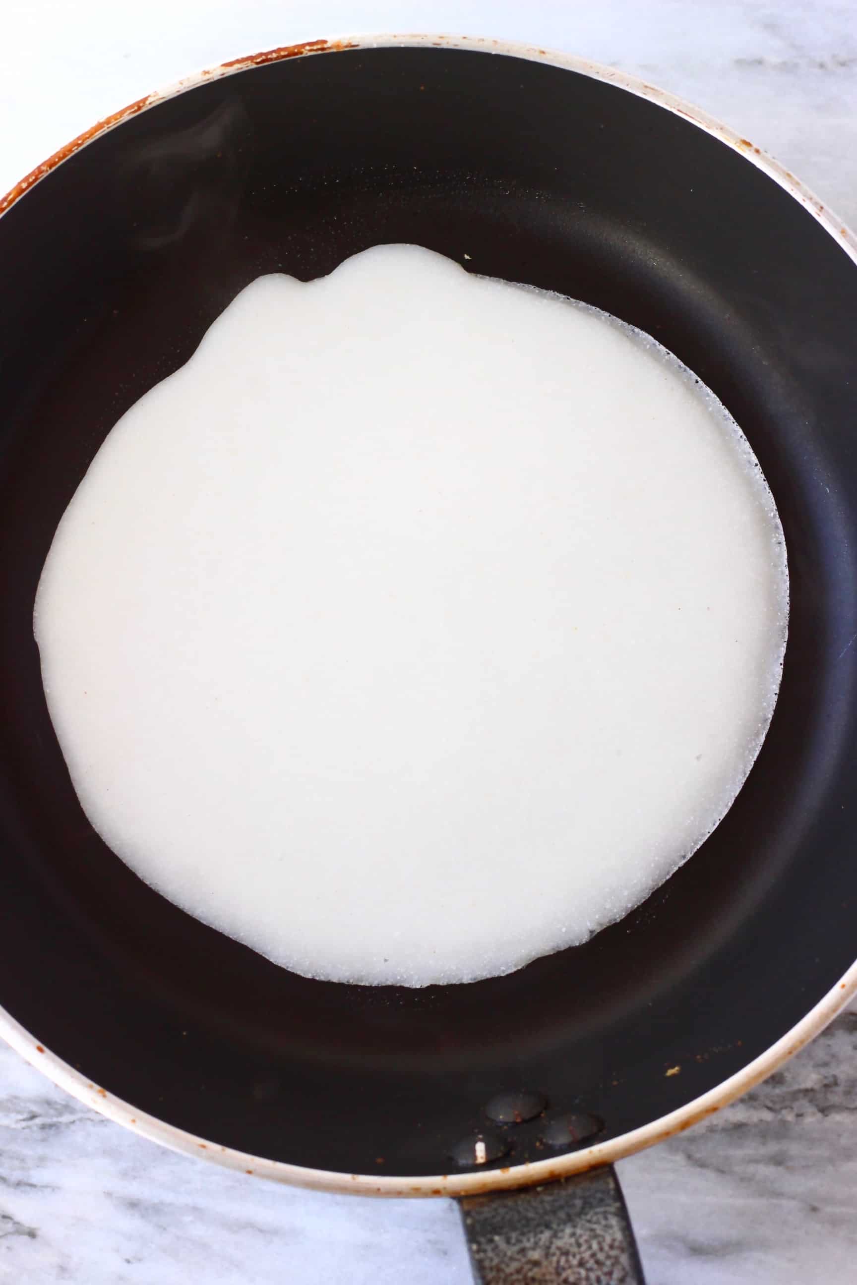 A round gluten-free vegan crepe being fried in a black frying pan 