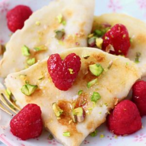 Three folded gluten-free vegan crepes on a white plate with pink flowers decorated with fresh raspberries and chopped pistachios with a gold fork against a marble background