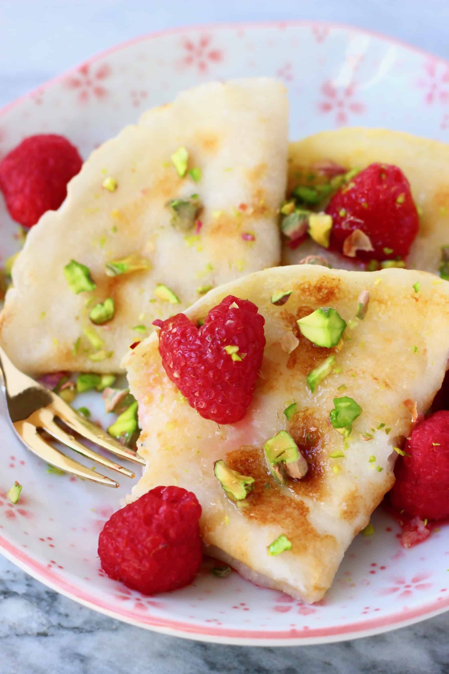 Three folded gluten-free vegan crepes on a white plate with pink flowers decorated with fresh raspberries and chopped pistachios with a gold fork against a marble background