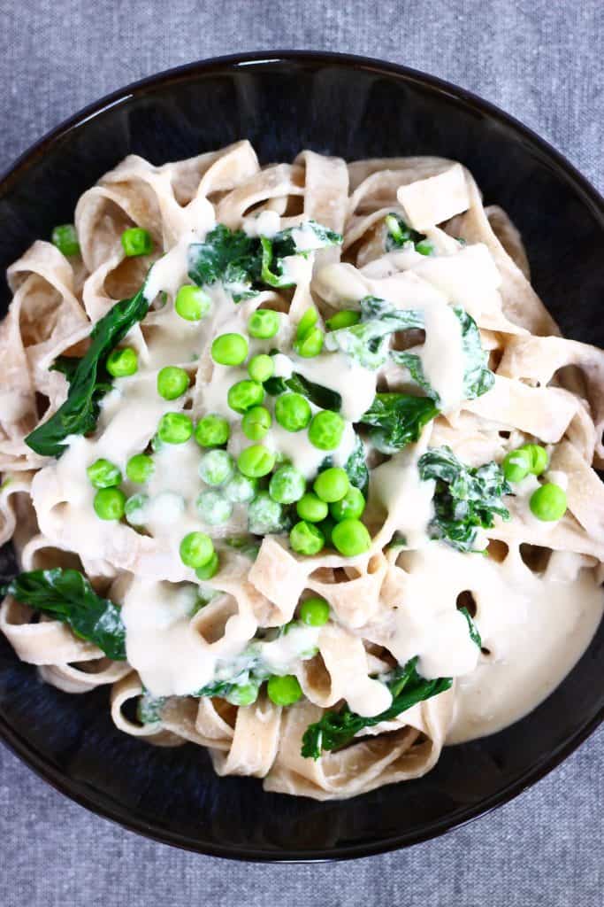 Tagliatelle in creamy cashew sauce with spinach and green peas in a black bowl against a grey background
