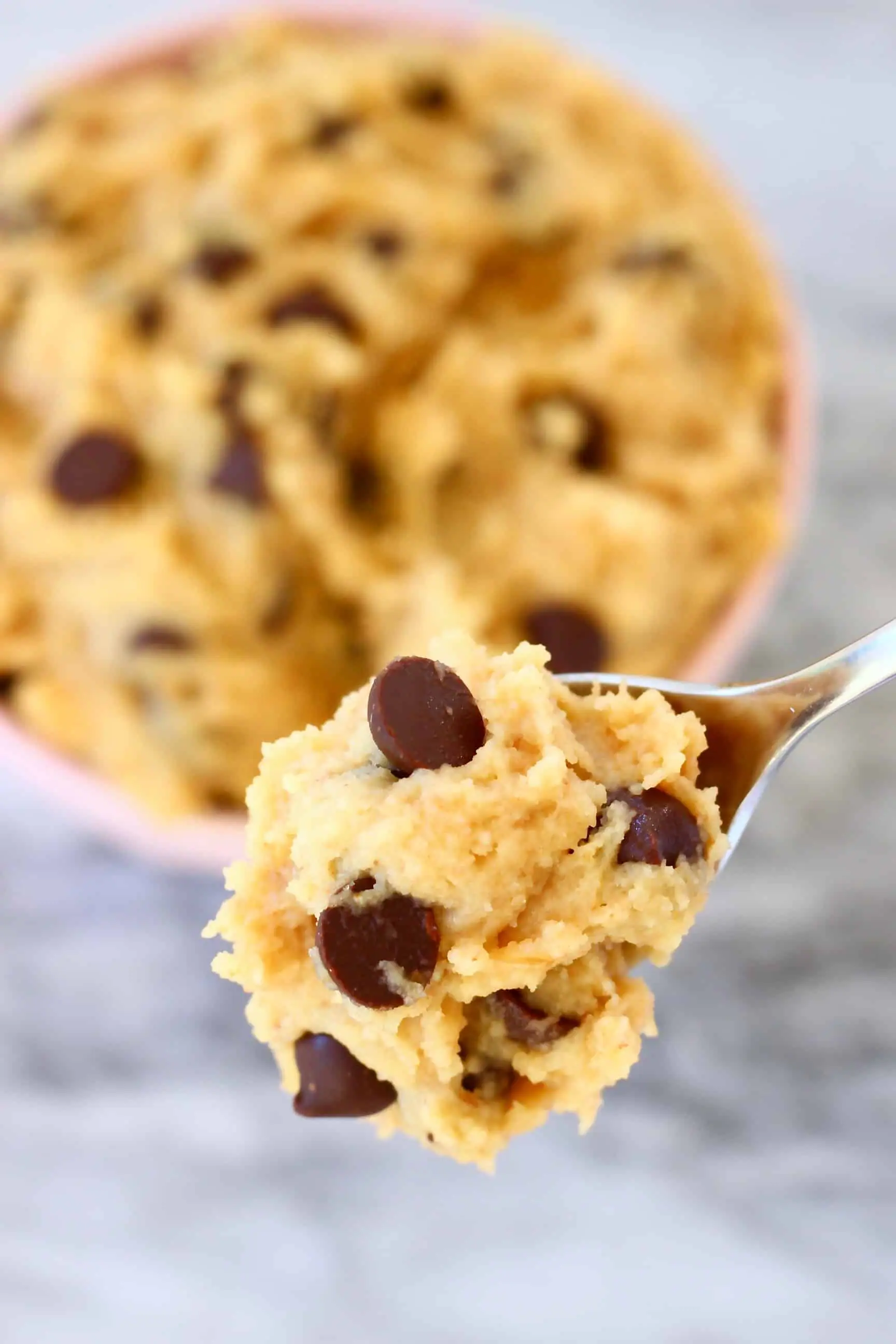 Vegan cookie dough with chocolate chips being lifted up with a spoon with a bowl of it in the background