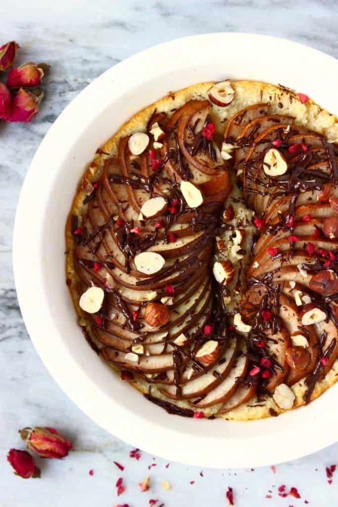 Photo of a pudding topped with sliced pears, chocolate and halved walnuts in a white pie dish against a marble background decorated with dried roses
