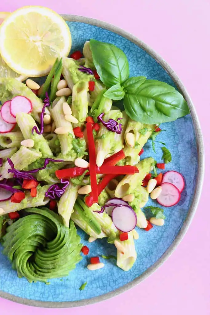 Penne pasta with avocado sauce decorated with colourful vegetables on a blue plate against a pink background