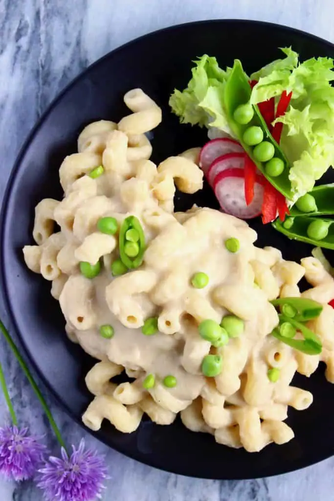 Photo of mac and cheese with green peas on a black plate with a side salad against a marble background