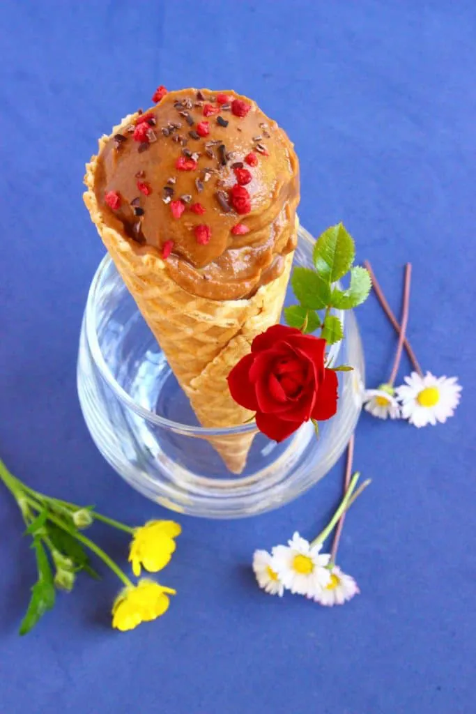 A glass with an ice cream cone filled with chocolate ice cream sprinkled with freeze-dried raspberries and cacao nibs against a bright blue background decorated with flowers