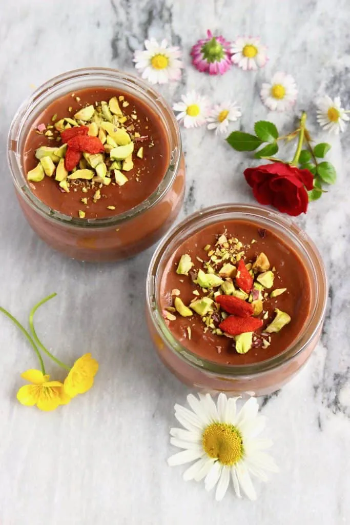 Two glass pots filled with chocolate pudding sprinkled with chopped pistachios and goji berries on a marble surface decorated with flowers