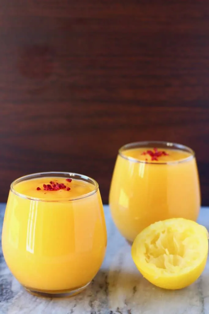 Two glasses filled with bright yellow lemon curd topped with freeze-dried raspberries on a marble surface with half a squeezed lemon against a dark brown background