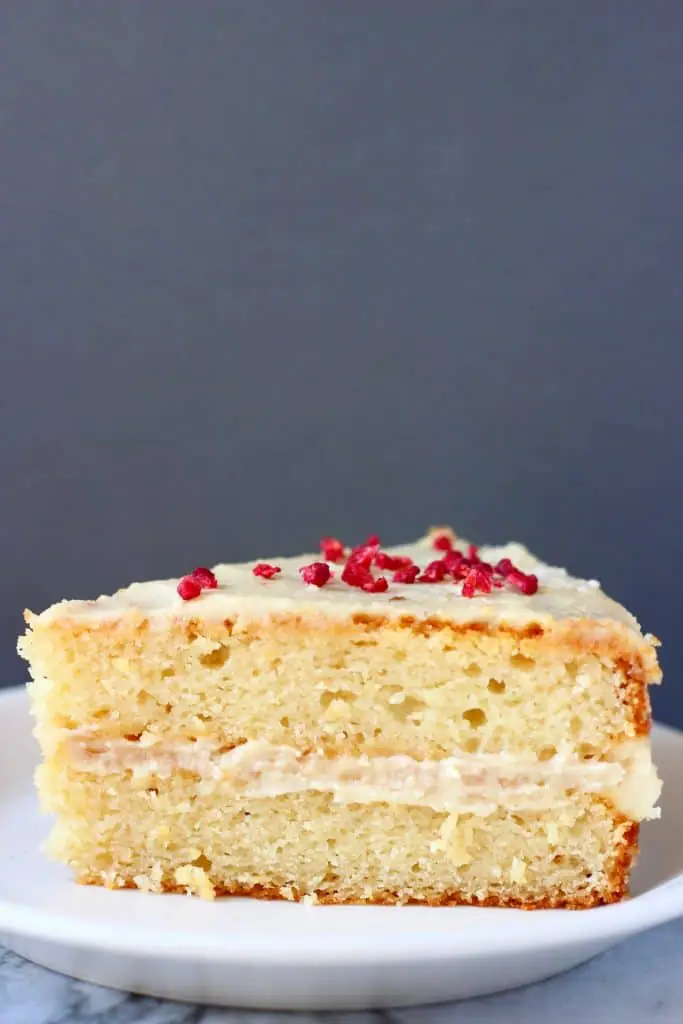 A slice of yellow sponge cake with white buttercream frosting sprinkled with freeze-dried raspberries on a white plate against a grey background