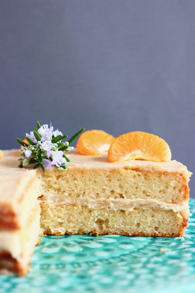 A sliced orange sponge cake with white buttercream frosting topped with clementine slices on a green cake stand against a grey background