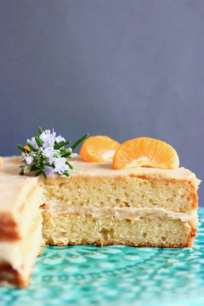 A sliced orange sponge cake with white buttercream frosting topped with clementine slices on a green cake stand against a grey background
