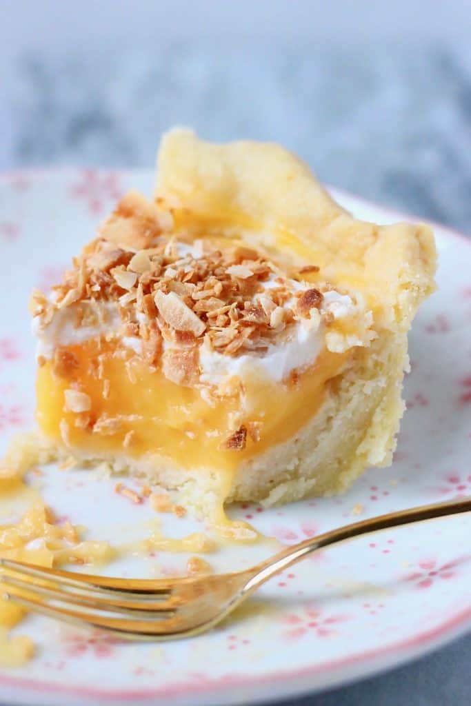 A slice of pie filled with lemon curd topped with cream and toasted coconut on a white plate with pink flowers with a small gold fork