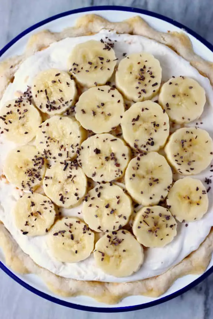 A pie crust filled with white cream and topped with banana slices and cacao nibs in a white pie dish with a dark blue rim against a marble background