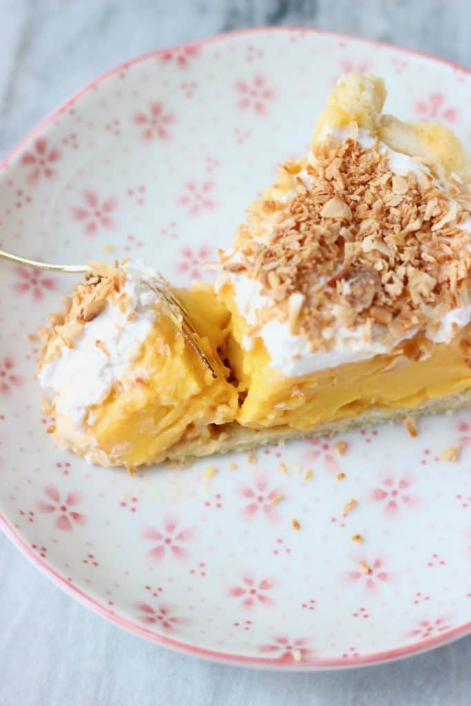 A slice of pie with yellow custard topped with white cream and toasted coconut on a white plate with pink flowers