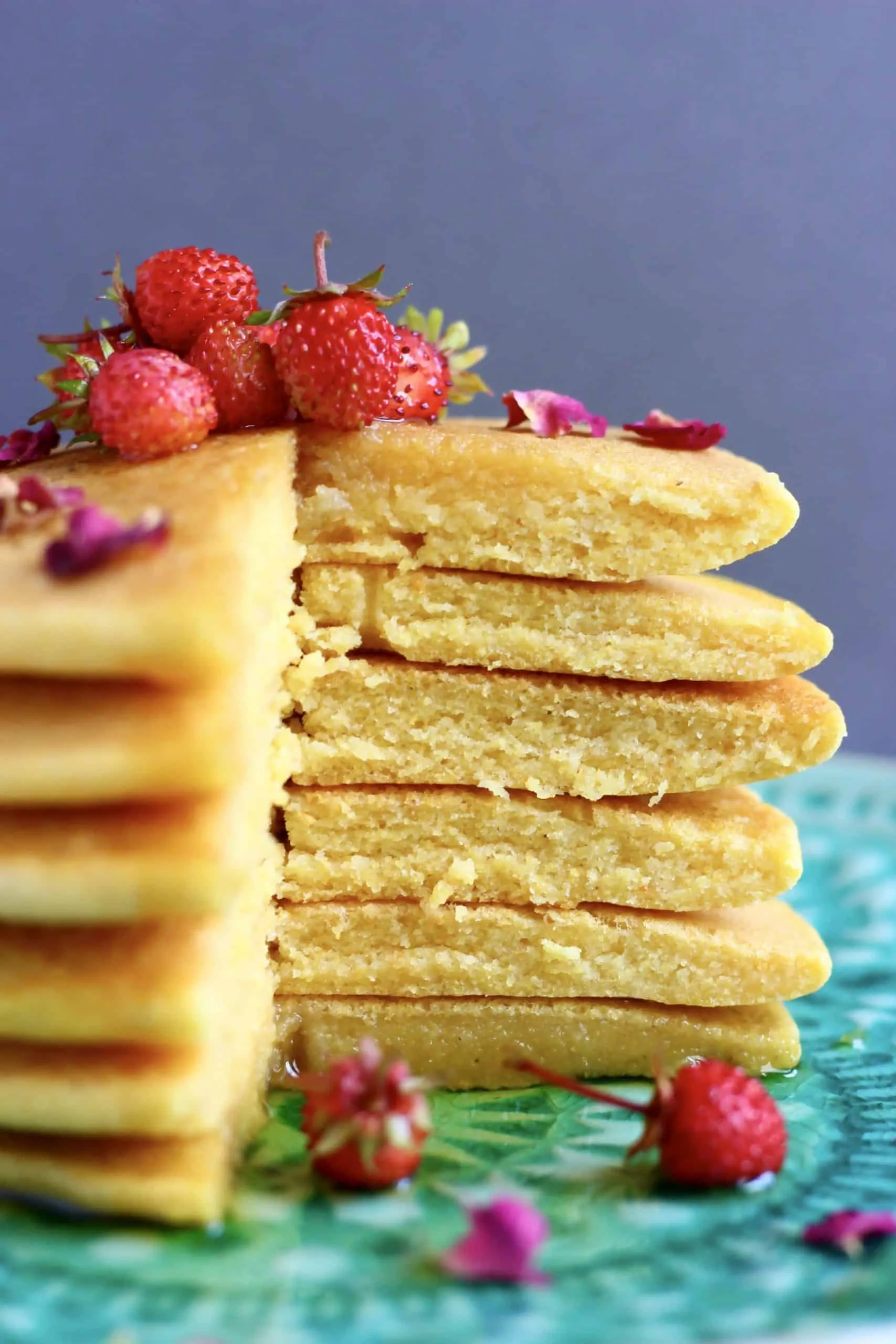 A stack of cornmeal pancakes with a slice cut out of them topped with rose petals and small strawberries on a green plate against a grey background