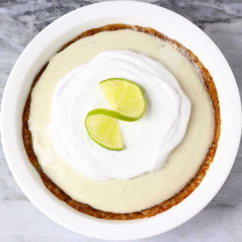 A brown pie crust filled with light green cream topped with white cream and a slice of lime in a white pie dish against a marble background