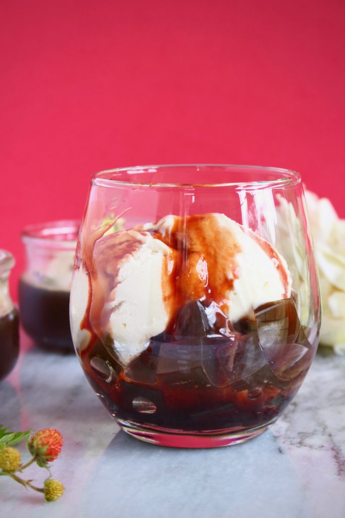 A glass filled with cubes of brown coffee jelly, two scoops of white ice cream and drizzled with a brown syrup on a marble surface against a bright pink background