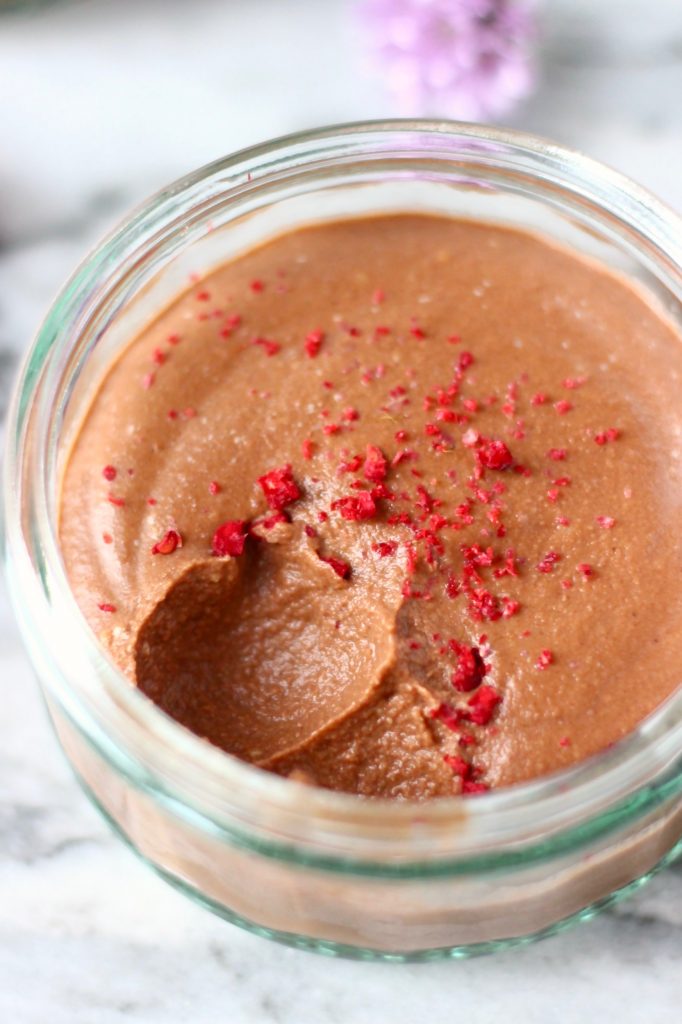 Photo of chocolate mousse in a glass ramekin topped with freeze-dried raspberries with a spoonful taken out of it against a marble background