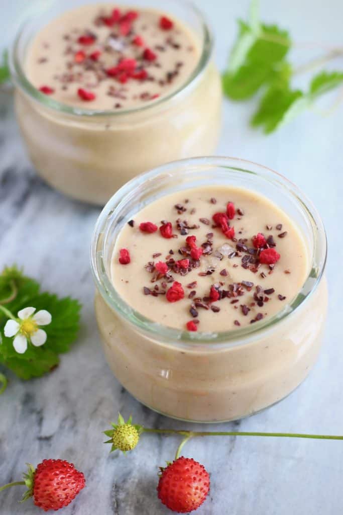 Two glass pots filled with beige-coloured pudding sprinkled with freeze-dried raspberries and cacao nibs against a marble background with mini strawberries
