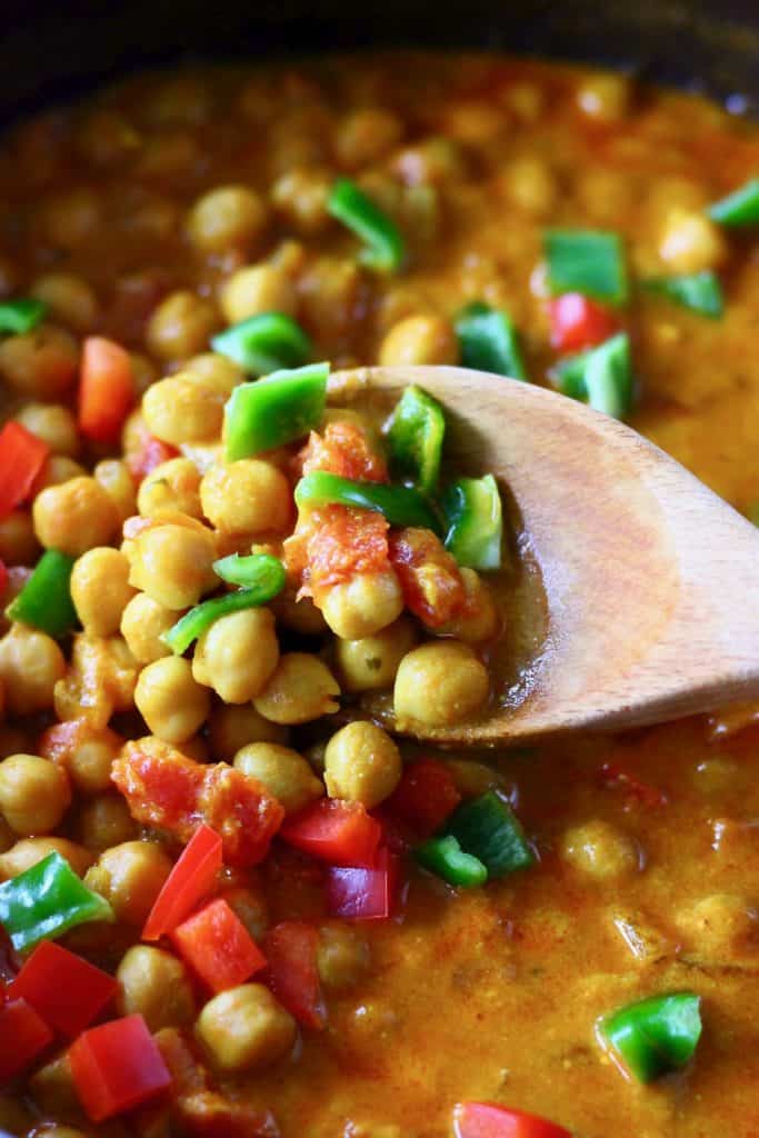 Red curry sauce with chickpeas, red peppers and green peppers in a black pan with a wooden spoon holding up a mouthful of curry
