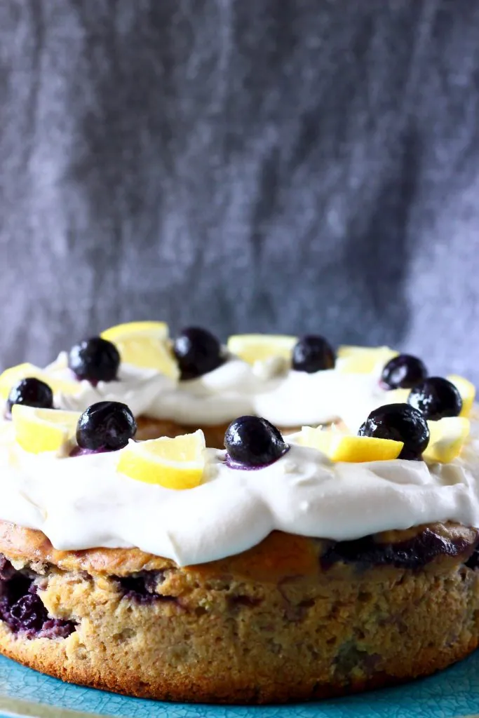 A round bundt cake topped with white creamy frosting, lemon slices and fresh blueberries against a grey background
