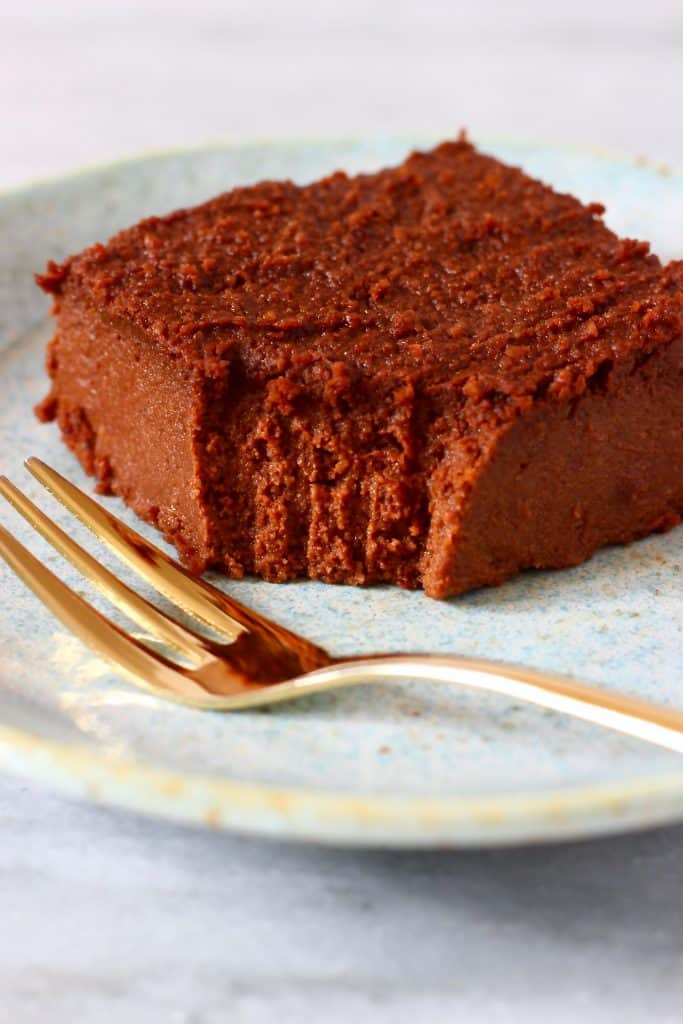 Photo of a square chocolate brownie with a bite taken out of it on a blue plate with a gold fork