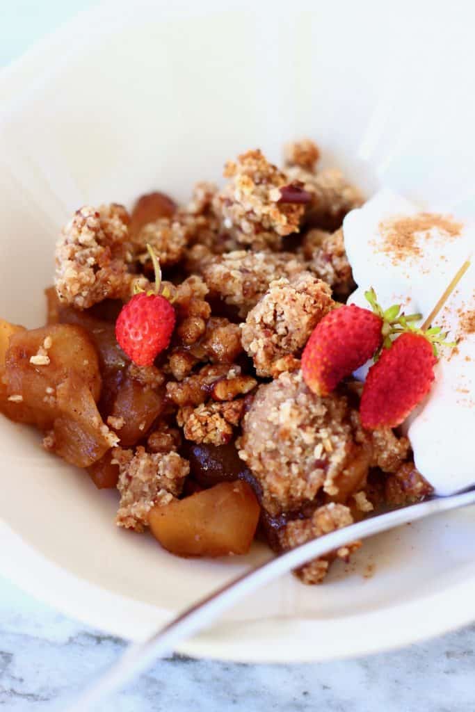 Photo of a portion of apple crumble and coconut yogurt in a white bowl topped with mini strawberries