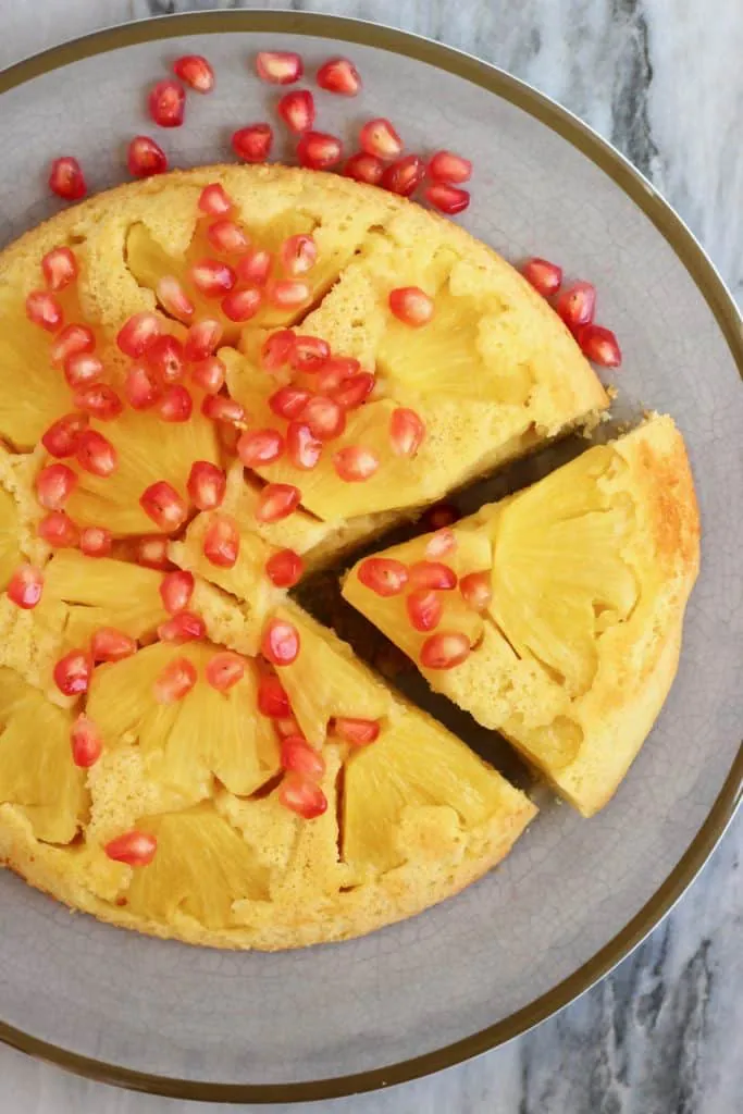 A pineapple upside down cake with a slice cut out of it sprinkled with pomegranate seeds on a grey plate against a marble background