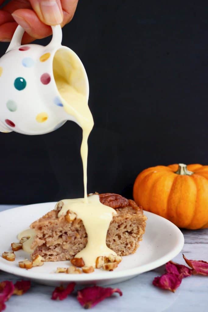 Photo of a small spotted jug of custard being poured over a slice of cake on a white plate against a black background