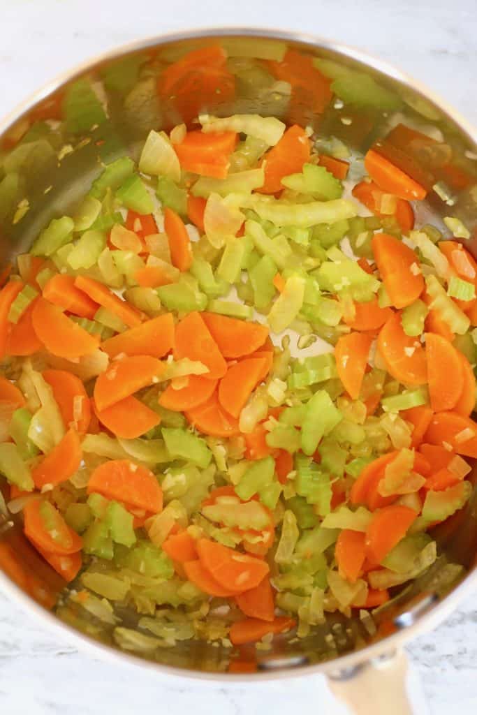 Onion, carrot and celery being fried in a silver pan against a marble background