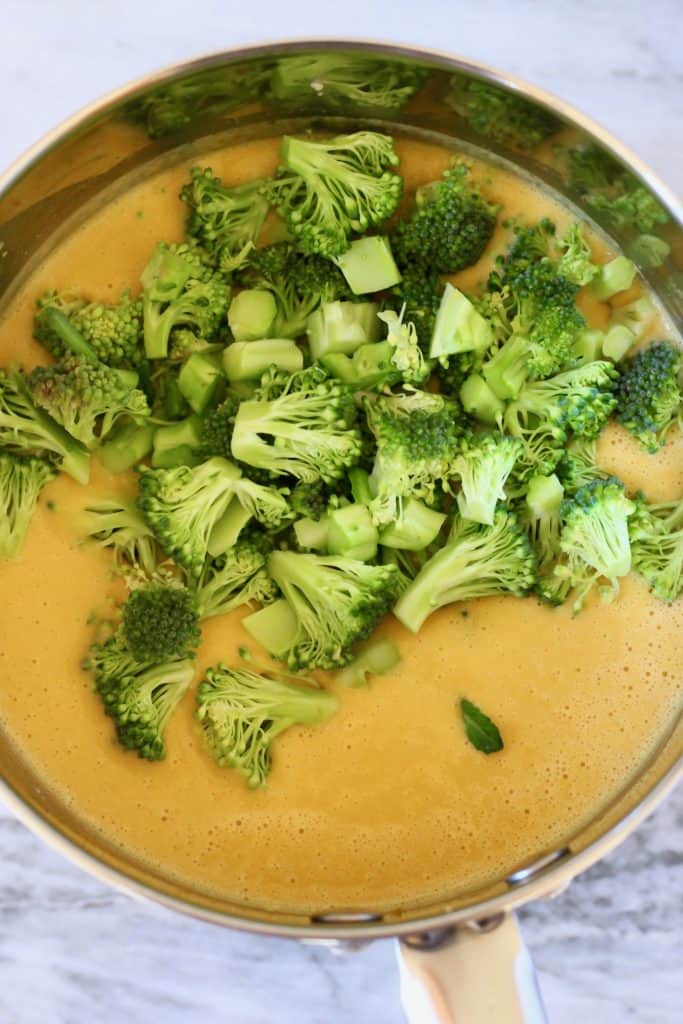 Yellow soup with raw broccoli in a silver saucepan against a marble background