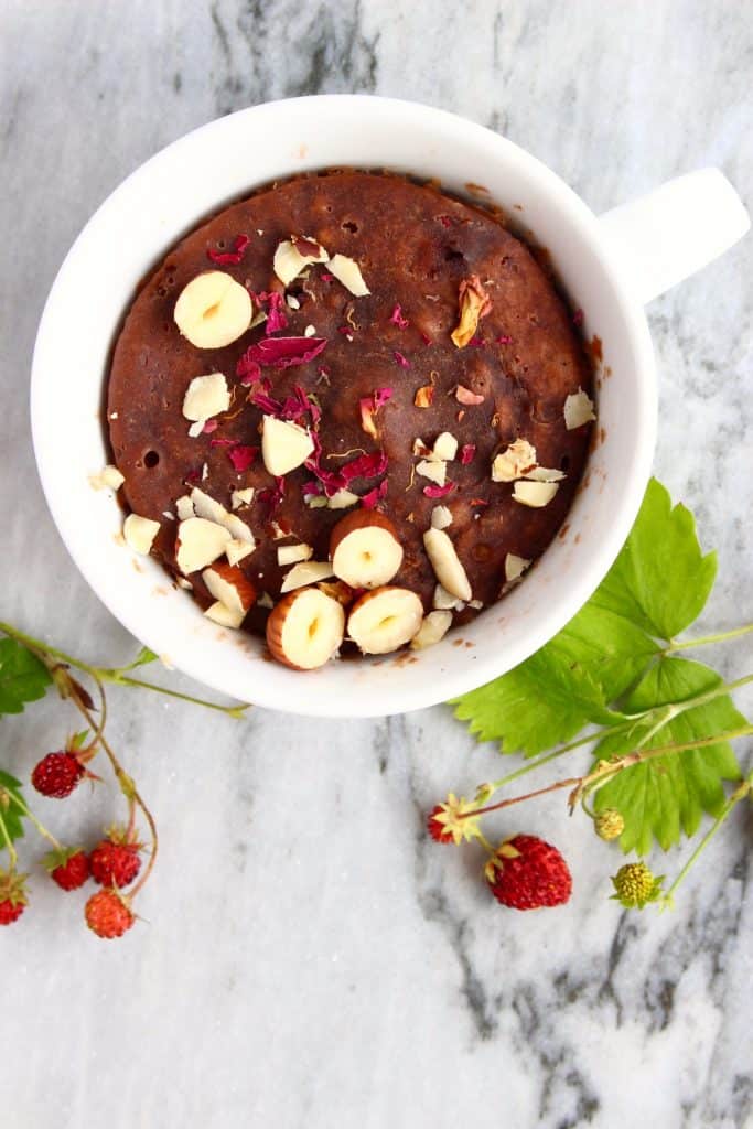 A chocolate cake in a white mug topped with chopped hazelnuts on a marble background decorated with wild strawberries