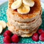 Stack of five banana pancakes topped with banana slices on top on a green plate with raspberries against a marble background