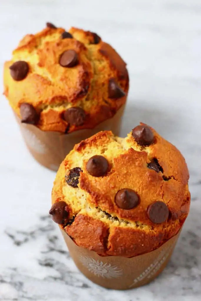 Two chocolate chip muffins in brown muffin cases against a marble background
