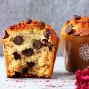 Two chocolate chip muffins in brown muffin cases on a marble slab against a grey background