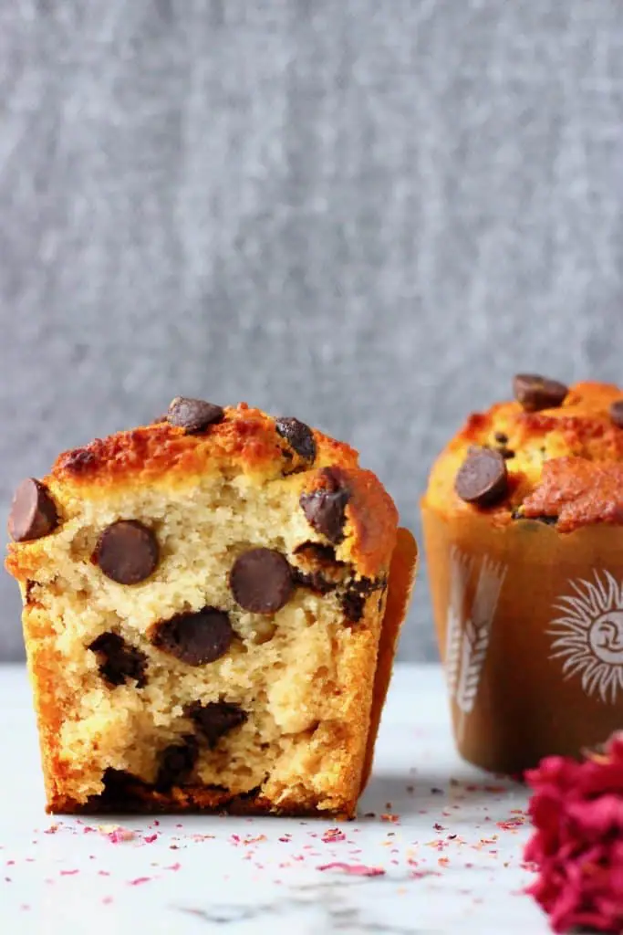Two chocolate chip muffins in brown muffin cases on a marble slab against a grey background