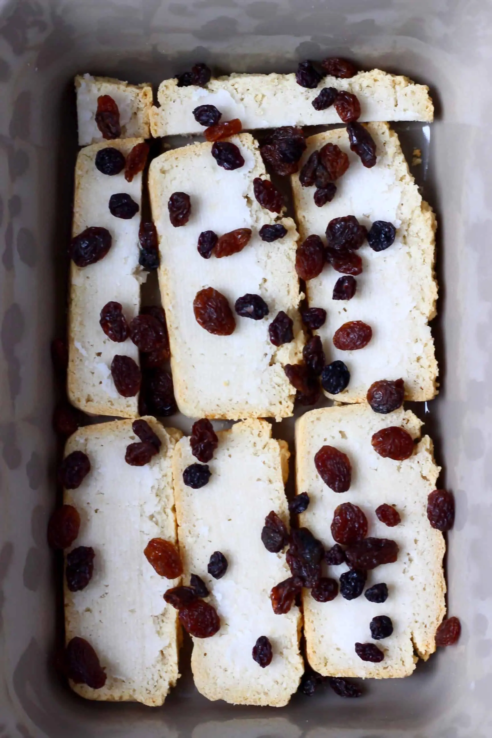 Slices of bread spread with coconut oil and topped with raisins in a grey rectangular baking dish
