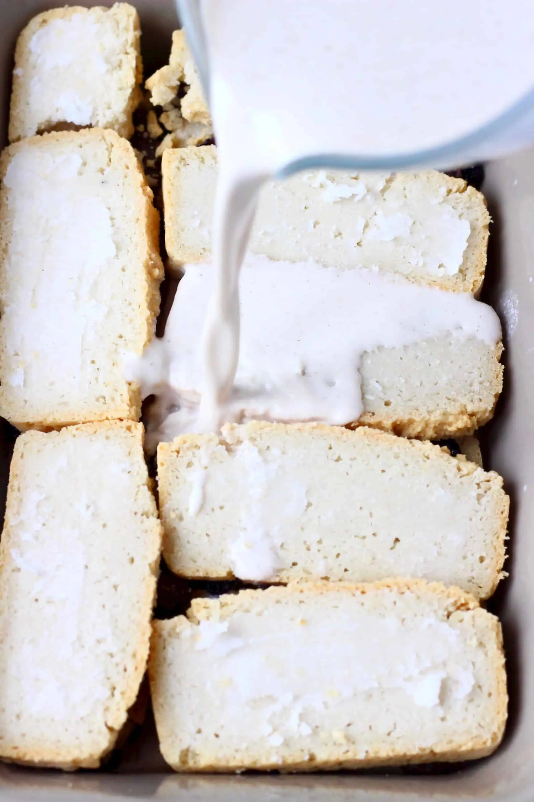 Slices of bread in a grey rectangular baking dish with a white cream being poured over the top with a glass jug