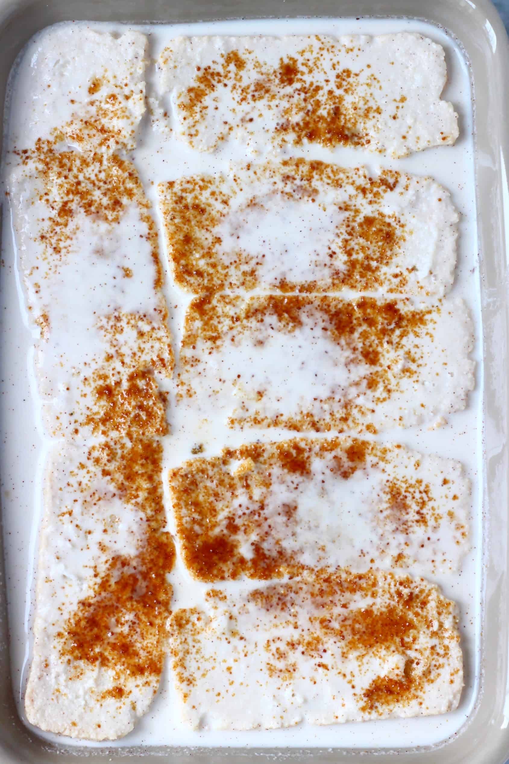 Slices of bread in a grey rectangular baking dish covered in white cream and sprinkled with brown coconut sugar