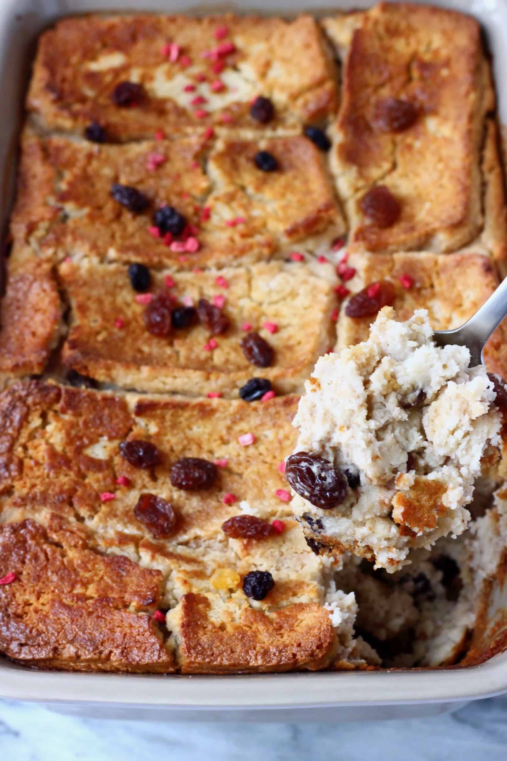 Vegan bread pudding in a grey rectangular baking dish sprinkled with raisins with a mouthful being held up with a spoon
