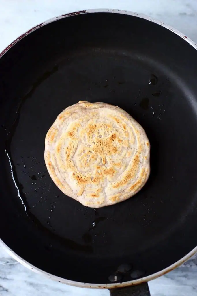 A buckwheat pancake being cooked on a black frying pan against a marble background
