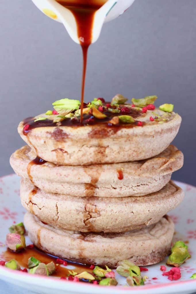 A stack of four buckwheat pancakes on a white plate decorated with chopped pistachios and freeze-dried raspberries with a white jug pouring syrup over it against a grey background